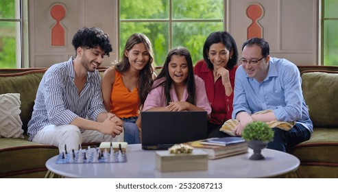 Indian extended family group use laptop watch television film TV feels happy spend time together indoor house. Young adult girl boy sitting cozy sofa couch look movie drama show enjoy having fun joy - Powered by Shutterstock