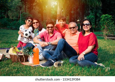 Indian Family Enjoying Picnic - Multi Generation Of Asian Family Sitting Over Lawn Or Green Grass In Park With Fruit Basket, Mat And Drinks. Selective Focus