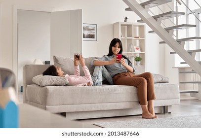 Indian Family Mother With Teen Daughter Using Phones Devices At Home. Young Mom With Teenage Child Holding Cellphones Relaxing Together On Couch In Living Room. Social Media Technology Addiction.