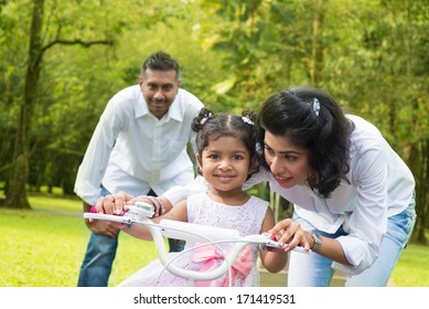Indian Family Outdoor Activity. Asian Parent Teaching Child To Ride A Bike At The Park In The Morning.