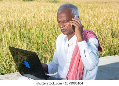 Indian Farmer Talking On Mobile Phone While Busy Looking Into Laptop Near The Agriculture Farmland - Concept Of Farmer Using Technology, Internet In Rural India