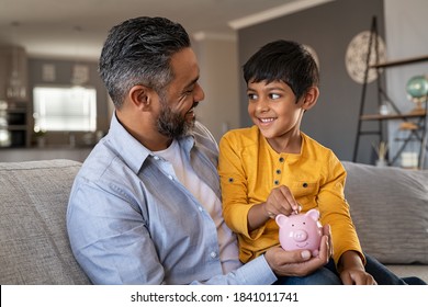 Indian father and smiling son putting coin into piggy bank. Smiling boy sitting on father lap saving money in piggybank. Middle eastern dad teaching son to save money while putting coin in piggy bank.