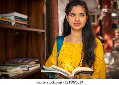 Indian Female College Student Reading A Book By Standing Near An Outdoor Library Bookcase At College Campus. She Is Looking Away And Thinking Something While Reading A Book. 