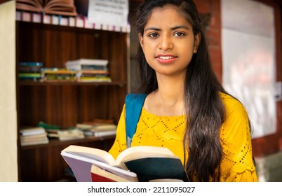 Indian Female College Student Reading A Book By Standing Near Outdoor Library Bookcase At College Campus And Looking At The Camera With Confident Smile.  