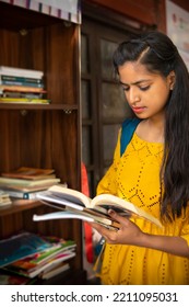 Indian Female College Student Reading A Book By Standing Near An Outdoor Library Bookcase At College Campus.  