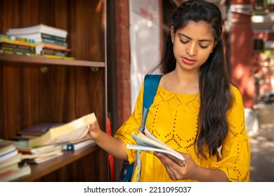 Indian Female College Student Reading A Book By Standing Near An Outdoor Library Bookcase At College Campus.  