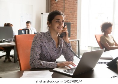 Indian female company member talking with corporate client customer on landline phone sitting in shared office with diverse colleagues typing on notebook, professional workers busy workday hr concept - Powered by Shutterstock