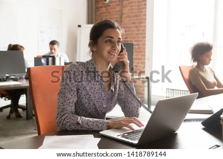 Indian female company member talking with corporate client customer on landline phone sitting in shared office with diverse colleagues typing on notebook, professional workers busy workday hr concept