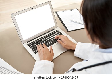 Indian female doctor wear white coat typing using laptop computer mock up white screen browsing internet sit at work desk. Healthcare medical e health website tech concept. Close up over shoulder view