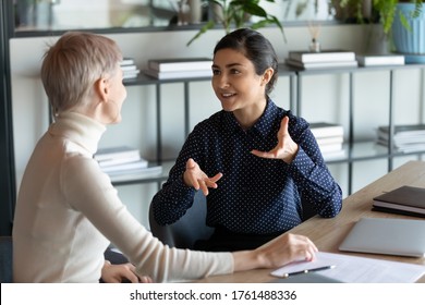 Indian female employee talking with Caucasian mate seated at workplace desk expresses her opinion on current issue, proposes solution to problem, share thoughts while working on common project concept