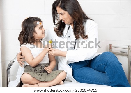 Indian female general practitioner doctor give lollipop to little girl patient after treatment or checkup at hospital.