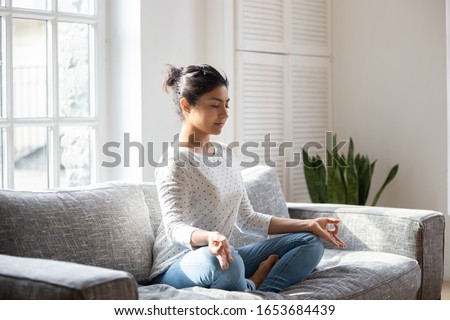 Indian female sitting on couch lotus pose put hands on lap folded fingers closed eyes enjoy meditation do yoga exercise at home. No stress, healthy habit, mindfulness lifestyle, anxiety relief concept