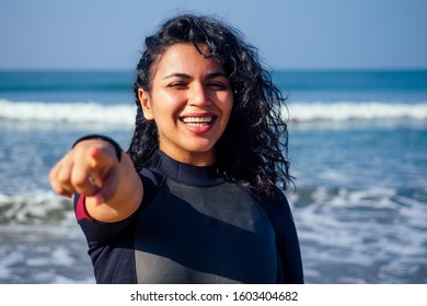 Indian Female Surfer Dressed In Black Wet Suit Is Standing Near Waters Edge With One Hand Pointing Finger At Camera