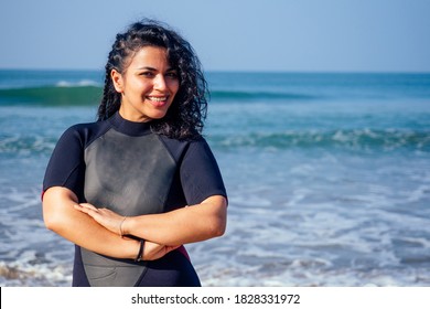 Indian Female Surfer Dressed In Black Wet Suit Is Standing Near Water With Crossed Hands