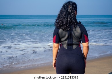 Indian Female Surfer Dressed In Black Wet Suit Looking At Sea , Back