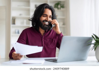 Indian freelancer man discussing work on phone call while reviewing documents and using laptop, smiling eastern male working in bright, contemporary home office setting, enjoying remote career - Powered by Shutterstock