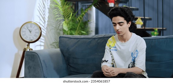 Indian frustrated teenager boy sitting on sofa feeling bad for heartbroken relationship at indoor home. sad teen age one worried alone man suffering fear tension regret mistake uncertain future