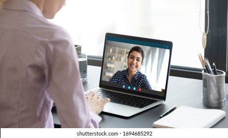 Indian girl communicate with friend on-line by video call, pc screen view over female shoulder. Mental health expert online therapy, colleagues work on common project use videoconferencing app concept