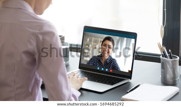Indian girl communicate with friend on-line by\
video call, pc screen view over female shoulder. Mental health\
expert online therapy, colleagues work on common project use\
videoconferencing app\
concept