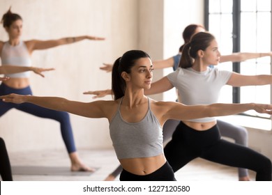 Indian girl and diverse group of young people practicing yoga lesson doing Warrior II exercise, Virabhadrasana 2 pose, working out, indoor close up, female students training at sport club or studio