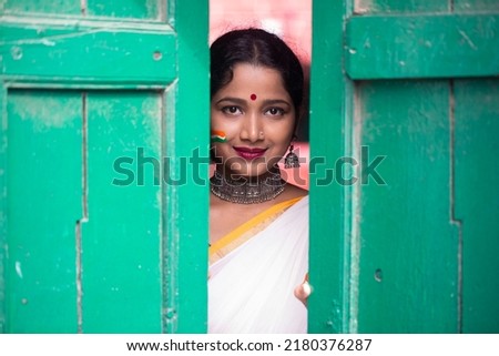 Indian girl face painted with Indian flag colors is peeping in the door. Portrait of a  girl is looking from the door. Beautiful eye of a child on black background. Republic or Independence day India