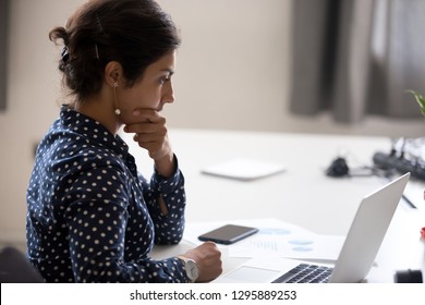 Indian girl sitting at desk near computer cogitating thinking making important decision at workplace. Concentrated serious office worker millennial woman analysing results feels doubts and feel unsure