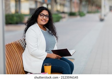 An Indian girl is sitting on a bench, reading a book and drinking coffee.