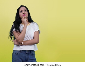 Indian girl thinking with finger on chin looking at left side with copy space. Beautiful Indian girl or Young south Asian woman thinking something deep isolated on Yellow background.