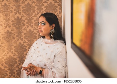 An Indian girl in white dress stands next to wall while as she is lost in thought. Bride wearing beautiful make up and traditional jewellery looking away.