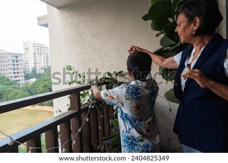 An Indian grandmother, wearing a white shirt and blue sleeveless jacket with jeans, helps her grandson put up a string of fairylights on the bannister in the balcony of their home in Mumbai, India.