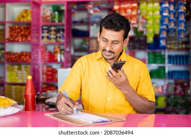 Indian groceries business man noting orders while talking with customer on mobile phone at kirana shop - concept of distance shopping, home delivery service.