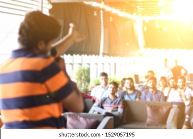 Indian Guy Doing Stand Up Comedy In Front Of Audience On Stage. Youth Audience Watching Performance In India. Blurred Event Photography.