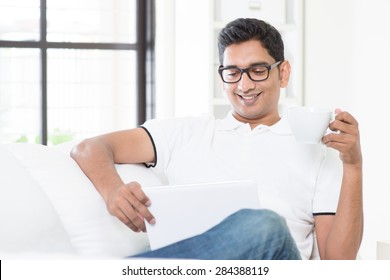 Indian Guy Enjoying A Cup Of Hot Tea While Using Digital Pc Tablet At Home. Asian Man Relaxed And Sitting On Sofa Indoor. Handsome Male Model.