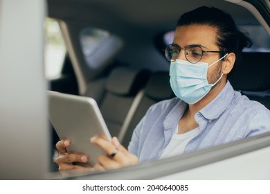 Indian Guy In Face Mask Using Digital Tablet While Taking Taxi, Copy Space. Middle-eastern Man Having Car Trip, Sitting On Back Seat And Reading Blog On Pad. Transportation Service During Lockdown