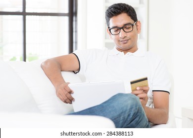 Indian Guy Hand Holding Credit Card, Enjoying Internet Online Shopping Using Digital Computer Tablet At Home. Asian Man Relaxed And Sitting On Sofa Indoor. Handsome Male Model.