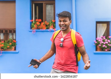 Indian Guy Smiling To The Camera Over Blue Background. Young Man Wearing Yellow Backpack Outdoors. Lifestyle Concept.