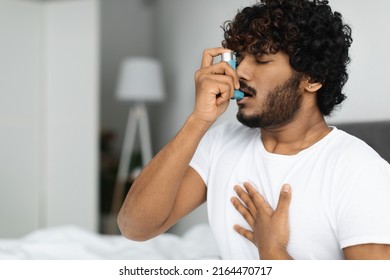 Indian guy suffering from asthma, using inhaler in bed, side view, copy space. Young hindu man woke up with asthma attack, touching chest and using nebulizer, bedroom interior