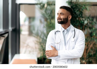 Indian handsome confident general medicine doctor, stands in a clinic, dressed in medical clothes with a stethoscope around his neck, looks away, smiling friendly. Doctor of the highest qualification
