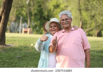 Indian happy senior couple showing thumb in garden and looking in front of camera