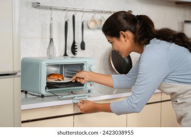 Indian happy smiling woman cooking burger by placing it on microwave oven at kitchen - concept of healthy eating, domestic lifestyle and technology