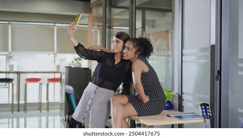 Indian happy teen girl hold mobile phone look front cam take selfie together indoor job work place. Smiling young adult gen z corporate woman make content video shoot during office break spend time - Powered by Shutterstock