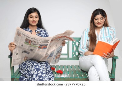 Indian happy women reading newspaper and story book while sitting on bench isolated on grey studio background. - Powered by Shutterstock