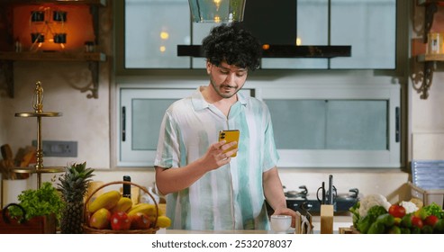 Indian happy young adult gen z handsome man standing use mobile phone typing message to friend chatting online look screen cam watch social media funny video enjoy day indoor modern kitchen home - Powered by Shutterstock