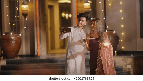 Indian Hindu happy gen z couple pair standing hold mobile phone take cute funny pose selfie outdoor home. Smiling young adult husband wife enjoy Diwali festival celebrate festive season outside house - Powered by Shutterstock