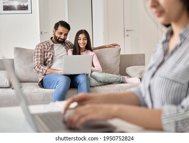 Indian Husband Spending Time With Teen Kid Daughter While Wife Working Online At Home. Busy Parent Mom Using Laptop Computer Sitting At Table In Home Office With Dad And Child In The Background.