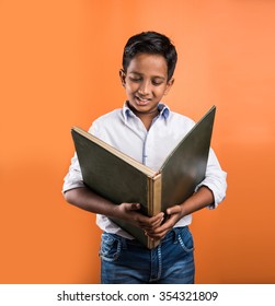 Indian Kid Enjoying Reading Or Holding Big Book, Standing Isolated Over Orange Background
