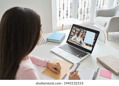 Indian kid girl having virtual class with online teacher distance learning at home. Latin teen school child student studying remote homeschool lesson on laptop computer with tutor. Over shoulder view