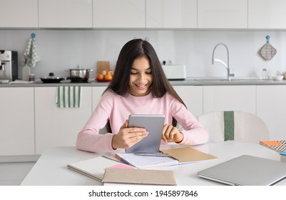 Indian Kid School Girl Distance Learning Online At Home Using Digital Tablet. Happy Child Teen Student Remote Studying Holding Pad Computer Homeschooling Doing Homework Sitting At Kitchen Table.