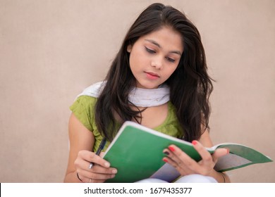 Indian Late Teen Girl Student Sitting Near Wall And Doing Study. She Is Reading And Writing On Book In Outdoor At Day Time. She Is Wearing Traditional Dress 