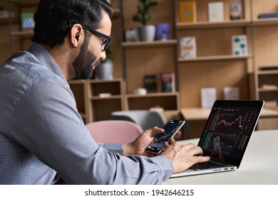 Indian Latin Business Man Trader Investor Analyst Looking At Financial Stock Market Charts Rate Dynamic, Trading Data Indexes Growth Using Mobile Phone And Laptop For Cryptocurrency Digital Analytics.